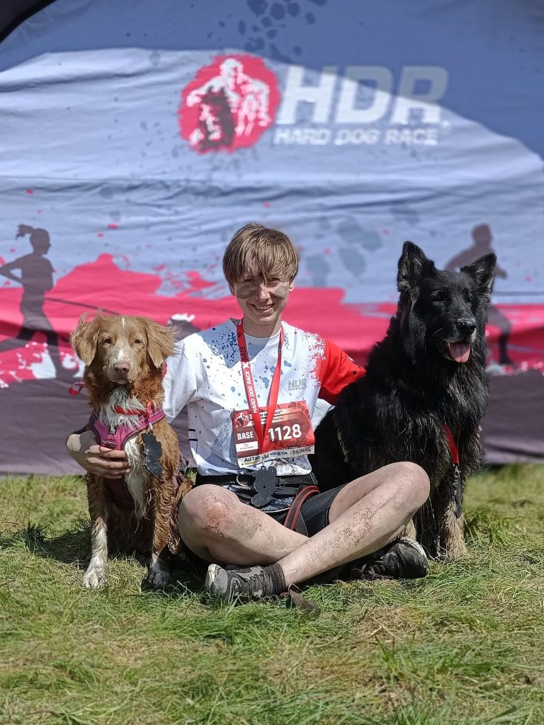 Ekipa po starcie w Hard Dog Race Base. Brudni od błota, zmęczeni, ale szczęśliwi: opiekun, toller i owczarek staroniemiecki pozują z medalami na tle ścianki zawodów.