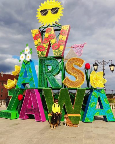 Kolorowy, kwiatowy napis "WARSZAWA" – popularna fotospot i instalacja miejska w stolicy Polski. Na zdjęciu widać osobę z dwoma psami pozującą na tle napisu ozdobionego słonecznikami, makami i słońcem w okularach. W tle widoczny jest plac na Starym Mieście.