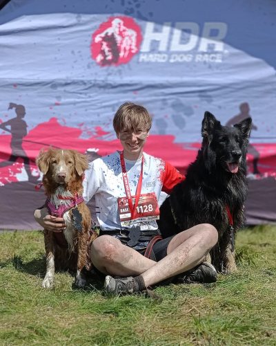 Ekipa po starcie w Hard Dog Race Base. Brudni od błota, zmęczeni, ale szczęśliwi: opiekun, toller i owczarek staroniemiecki pozują z medalami na tle ścianki zawodów.
