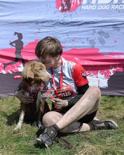 Uczestniczka zawodów Hard Dog Race Base całuje swojego tollera po biegu. Siedzą na trawie z pamiątkowymi medalami, ubrudzeni błotem, na tle banera zawodów.